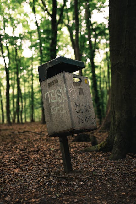 A grey metal public mailbox is situated in a forested area, attached to a single black pole embedded in the ground. The mailbox has a slightly weathered appearance with visible graffiti and stickers on its surface, including some black and yellow markings near the top. The postal slot is on the front panel, slightly off-center, with a small raised ledge on top serving as a cover. The surroundings consist of tall trees with rough, dark brown bark, and a forest floor covered in brown soil and scattered leaves, with dappled sunlight filtering through the dense canopy above. In the background, the blurred greenery of leaves and branches indicates a dense woodland environment. The scene suggests an unusual placement for a mailbox, possibly indicating an independent collection or public access point in a natural setting, aligning with the concept of alternative waste or rubbish handling in outdoor environments. The overall scene is quiet and natural, with no other objects or signs of urban infrastructure visible.
