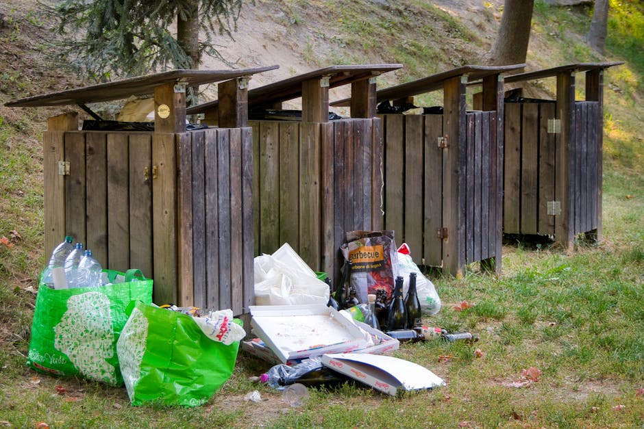 An outdoor scene shows four dark brown wooden rubbish bins with pitched roofs, arranged in a row on a grassy area with a slight incline and some trees in the background. In front of the bins, there is a collection of discarded waste, including several large green plastic bags filled with bottles and packaging, scattered around the base. A white pizza box with leftover food residues rests on the ground, alongside empty glass bottles, plastic bottles, and cardboard packaging, indicating informal rubbish disposal. The environment appears to be a park or natural area, with the waste deliberately placed near the bins but spilling onto the grass, suggesting a possible unauthorised disposal or an overflow from private rubbish collection. The scene highlights the importance of proper waste management, as managed by services like Waste Removal Belsize Park, to prevent environmental littering and promote clean public spaces related to local rubbish collection efforts.