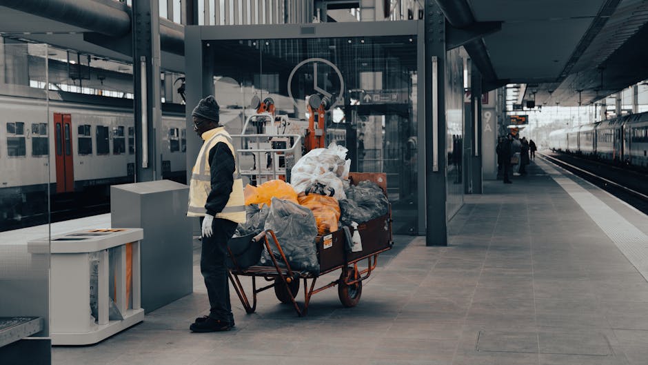 A man wearing a dark beanie, black jacket, and white gloves stands on a modern train station platform near a large trolley loaded with various types of rubbish, including plastic bags, cardboard, and miscellaneous waste. The trolley has a rust-colored metal frame and is filled with waste materials, some in black and orange plastic bags, and is positioned in front of a glass-enclosed area that appears to be part of the station's entrance or ticketing zone. In the background, a sleek train with red and white doors is parked alongside the platform, with additional passengers visible further down the platform. The station is characterized by contemporary architecture with steel beams, glass walls, and a clean, tiled floor. Overhead lighting provides natural and artificial illumination, contributing to a neutral and professional atmosphere. The scene suggests an instance of waste collection or disposal, possibly related to station maintenance or cleaning services by Waste Removal Belsize Park, illustrating an example of how waste is managed in a busy public transport environment. This detailed visual aligns with services related to rubbish removal and alternative waste handling at busy urban transit points.
