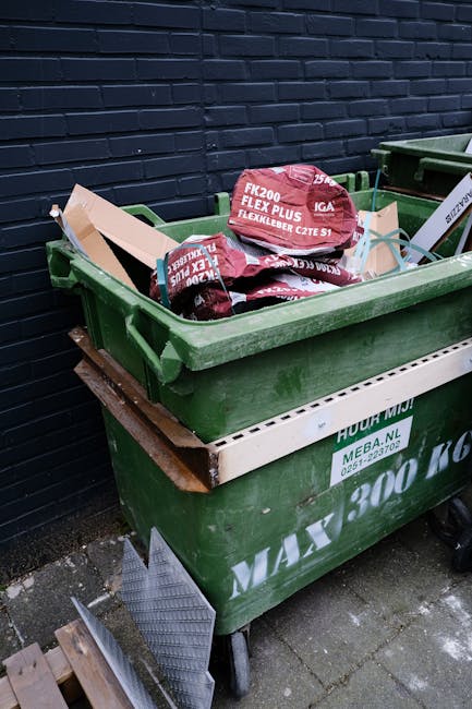 An outdoor scene shows four dark brown wooden rubbish bins with pitched roofs, arranged in a row on a grassy area with a slight incline and some trees in the background. In front of the bins, there is a collection of discarded waste, including several large green plastic bags filled with bottles and packaging, scattered around the base. A white pizza box with leftover food residues rests on the ground, alongside empty glass bottles, plastic bottles, and cardboard packaging, indicating informal rubbish disposal. The environment appears to be a park or natural area, with the waste deliberately placed near the bins but spilling onto the grass, suggesting a possible unauthorised disposal or an overflow from private rubbish collection. The scene highlights the importance of proper waste management, as managed by services like Waste Removal Belsize Park, to prevent environmental littering and promote clean public spaces related to local rubbish collection efforts.