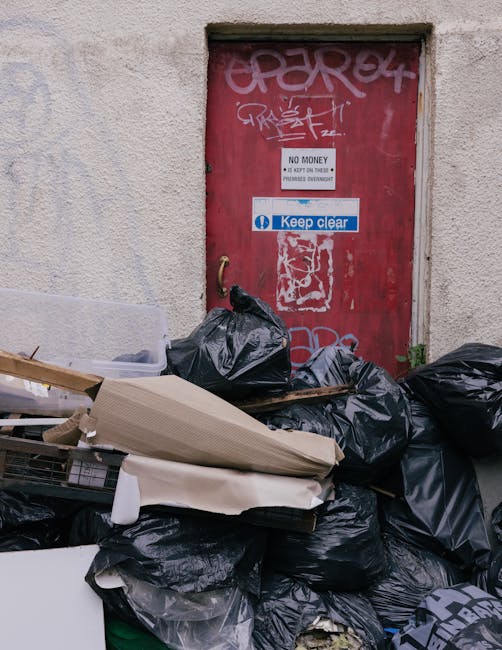 A large pile of black garbage bags, some torn open revealing waste, is stacked in front of a red metal door set into a light-colored, textured brick wall. The bags are overflowing onto the surrounding pavement, with a few loose items such as flattened cardboard boxes and discarded packaging visible among them. The red door features graffiti tags, a white sign indicating 'NO MONEY' and 'KEEP CLEAR' with blue lettering, and is secured with a brass handle on the left side. The scene suggests an area involving waste collection or disposal, potentially managed by a private rubbish removal service like Waste Removal Belsize Park, and indicates an on-site clearance of household or commercial rubbish in an urban environment.