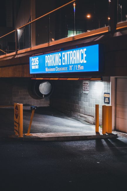 This image shows the entrance to a parking garage illuminated at night, with a large blue electronic sign displaying the gate number '235' and the words 'Parking Entrance' along with the maximum vehicle height restriction of '78 inches or 1.95 meters'. The entrance is bordered by yellow safety bollards and a yellow barrier arm, with a concrete driveway leading into the underground parking area. The surrounding environment includes a brick wall beneath the sign and a small warning sign on the wall, indicating safety instructions. The overall scene features artificial lighting with reflections on the wet pavement, emphasizing the structural details and safety measures in place for vehicle access. This setting illustrates typical private parking facilities which may require independent access and clearance, aligning with alternative waste management or service entry points often used by waste removal companies like Waste Removal Belsize Park for logistics and operational efficiency.