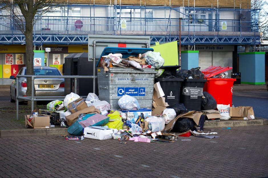 A large pile of black garbage bags, some torn open revealing waste, is stacked in front of a red metal door set into a light-colored, textured brick wall. The bags are overflowing onto the surrounding pavement, with a few loose items such as flattened cardboard boxes and discarded packaging visible among them. The red door features graffiti tags, a white sign indicating 'NO MONEY' and 'KEEP CLEAR' with blue lettering, and is secured with a brass handle on the left side. The scene suggests an area involving waste collection or disposal, potentially managed by a private rubbish removal service like Waste Removal Belsize Park, and indicates an on-site clearance of household or commercial rubbish in an urban environment.