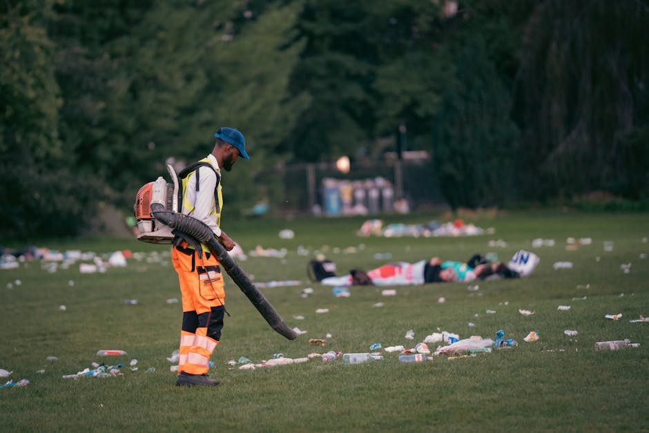 A person dressed in high-visibility orange trousers with reflective strips, a white safety vest, and a blue cap is using a leaf blower to clear litter from a grassy park area. The individual is standing in the foreground, with a backpack-style collection device attached to the leaf blower, which is directed downward to gather and disperse debris. The scene shows various types of rubbish, including plastic bottles, paper cups, and wrappers, scattered across the grass, with some being blown around by the equipment. In the background, there are two individuals lying on the ground, partially obscured by the scattered litter, with trees and bushes framing the scene and a distant row of recycling containers or waste disposal bins. The overall environment appears to be an outdoor urban park in early evening or late afternoon, highlighting the importance of waste management and park clean-up services provided by Waste Removal Belsize Park in supporting off-site rubbish collection and maintaining public spaces.