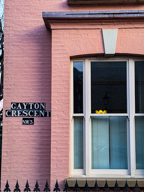 A close-up view of a pink brick building facade featuring a large, white-framed bay window with multiple panes. The window has a small ledge decorated with a yellow rubber duck and light blue curtains inside. To the left of the window, a black and white street sign indicates the corner of Gayton Crescent, NW3. The brickwork has a smooth texture with visible mortar joints, and the area is illuminated by natural daylight, casting soft shadows. Below the window, a black wrought iron fence with pointed finials runs horizontally across the image, suggesting the building's exterior. The overall scene portrays a residential setting, with the building's external features and street sign providing context for the local environment, typical of alternative waste handling and private rubbish collection services in the area managed by Waste Removal Belsize Park.